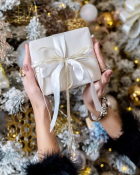 Female hands hold a gift box tied with a ribbon on the background of a Christmas tree, the concept of christmas and gifts