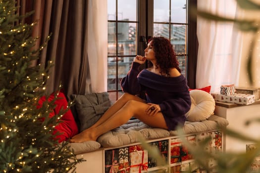 Woman christmas window. Beautiful curly woman in a purple sweater sitting on a windowsill near a Christmas tree, a room decorated for Christmas. Holiday at home