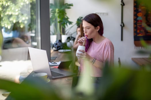 Side view of young businesswoman sitting at table in coffee shop. On table cup of coffee and laptop. In background white wall and window. Girl shopping online.