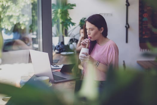 Side view of young businesswoman sitting at table in coffee shop. On table cup of coffee and laptop. In background white wall and window. Girl shopping online.