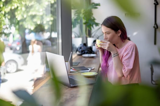 Side view of young businesswoman sitting at table in coffee shop. On table cup of coffee and laptop. In background white wall and window. Girl shopping online.