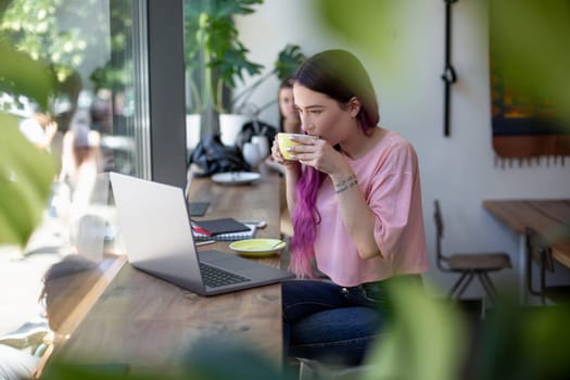 Side view of young businesswoman sitting at table in coffee shop. On table cup of coffee and laptop. In background white wall and window. Girl shopping online.