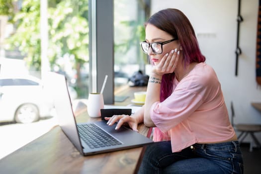 Side view of young businesswoman sitting at table in coffee shop. On table cup of coffee and laptop. In background white wall and window. Girl shopping online.