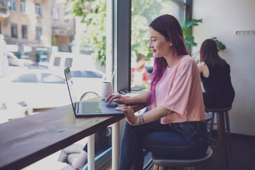 Side view of young businesswoman sitting at table in coffee shop. On table cup of coffee and laptop. In background white wall and window. Girl shopping online.