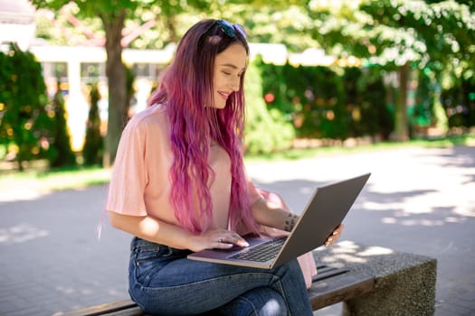 Young girl with pink hair is studying in the spring park, sitting on the wooden bench and browsing on her laptop