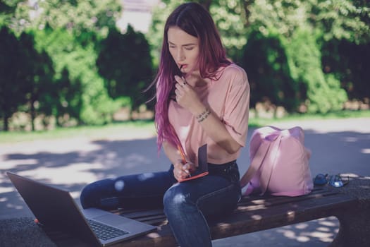 Woman writing in a notebook sitting on a wooden bench in the park. Girl working outdoors on portable computer, copy space. Technology, communication, freelance and remote working concept.