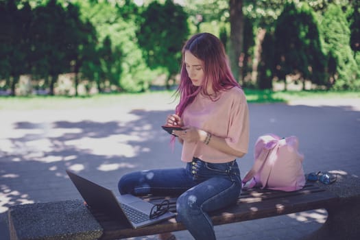 Woman writing in a notebook sitting on a wooden bench in the park. Girl working outdoors on portable computer, copy space. Technology, communication, freelance and remote working concept.