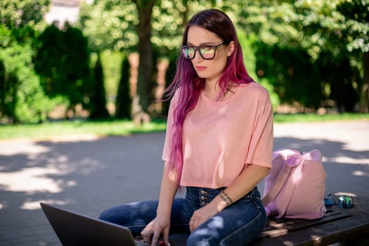 Young girl with pink hair is studying in the spring park, sitting on the wooden bench and browsing on her laptop