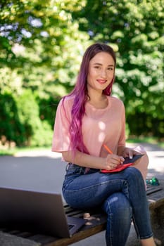 Woman writing in a notebook sitting on a wooden bench in the park. Girl working outdoors on portable computer, copy space. Technology, communication, freelance and remote working concept.