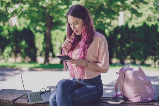 Woman writing in a notebook sitting on a wooden bench in the park. Girl working outdoors on portable computer, copy space. Technology, communication, freelance and remote working concept.