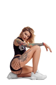 Attractive blond twerk lady with tattoed body and long curly hair is posing sitting on a floor, isolated on white background with copy space. Young girl wearing in a mesh black shorts, top and white sneakers. Booty dance in studio.