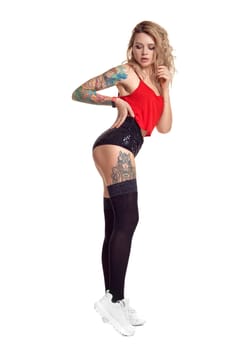 Sexy blond woman with tattoed body and long curly hair is looking down while posing isolated on white background with copy space. Young girl wearing in a black stockings and mini shorts, red top and white sneakers. Booty twerk dance in studio.