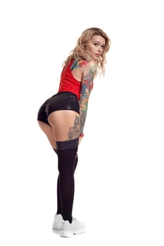 Charming blond maiden with tattoed body and long curly hair is looking a the camera while posing isolated on white background with copy space. Young woman wearing in a black stockings and mini shorts, red top and white sneakers. Booty twerk dance in studio.