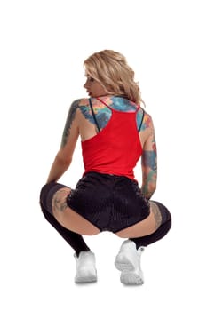 Charming blond lady with tattoed body and long curly hair is posing back, isolated on white background with copy space. Young woman wearing in a black stockings and mini shorts, red top and white sneakers. Booty twerk dance in studio.