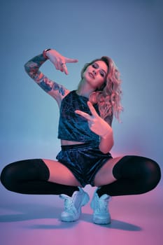 Beautiful young blonde tattoed girl in blue velour booty shorts and t-shirt, black stockings, squats in front of the camera in a confident pose. Pink and blue background