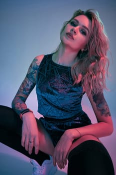 Beautiful young blonde tattoed girl in blue velour booty shorts and t-shirt, black stockings, squats in front of the camera in a confident pose. Pink and blue background
