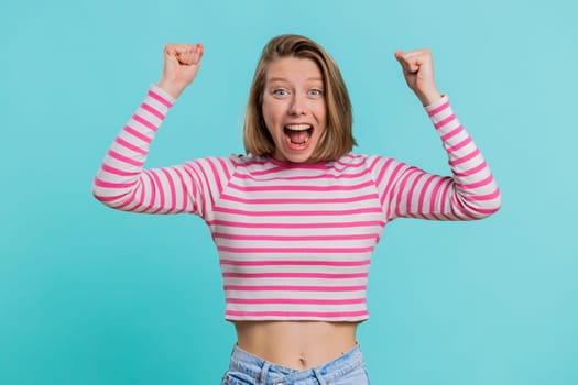 Happy joyful woman shouting, raising fists in gesture I did it, celebrating success, winning birthday, lottery jackpot goal achievemen, good news. Young girl in crop top on blue studio wall background