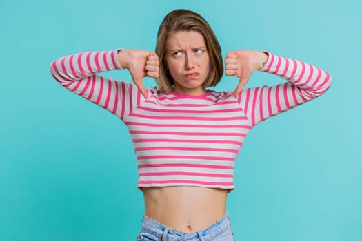 Dislike. Upset unhappy young woman showing thumbs down sign gesture, expressing discontent, disapproval, dissatisfied, dislike. Pretty girl in crop top. Indoors isolated on blue studio background