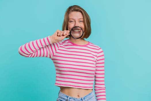 Young woman holding magnifier glass loupe on healthy white teeth, looking at camera with happy expression, showing funny silly face smiling mouth. Girl in crop top isolated on blue studio background