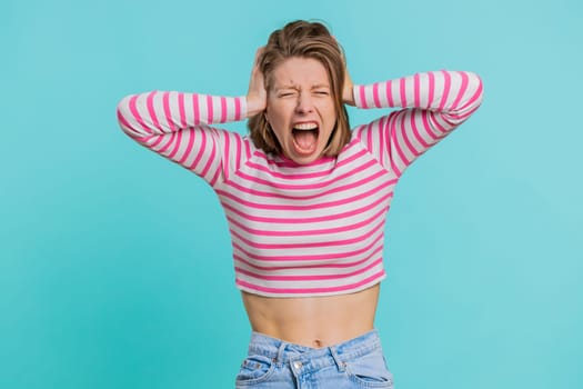 Young woman screams yell shout from stress tension problems feels horror hopelessness fear panic surprise shock expresses gestures rage. Pretty girl in crop top isolated on blue studio background