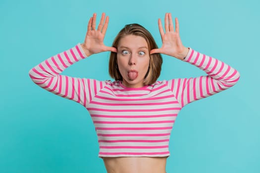 Funny comical playful young woman making silly facial expressions and grimacing, fooling around, showing tongue, idiotic expression. Pretty girl in crop top isolated alone on blue studio background