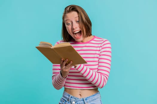 Young excited woman i reading funny interesting fairytale story book, laughing, leisure hobby, knowledge wisdom, education, learning study, wow. Girl in crop top isolated on blue studio background