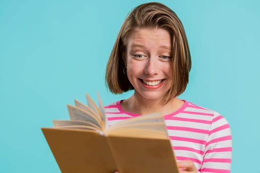 Young excited woman i reading funny interesting fairytale story book, laughing, leisure hobby, knowledge wisdom, education, learning study, wow. Girl in crop top isolated on blue studio background