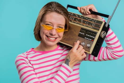 Woman in sunglasses using retro tape record player to listen music, disco dancing of favorite track, having fun, entertaining, fan of vintage technologies. Girl in crop top isolated on blue background