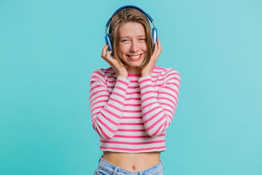 Happy woman in headphones listening music and dancing disco fooling around having fun expressive gesticulating hands relaxing on party. Pretty girl in sunglasses isolated on studio blue background