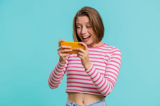 Worried funny addicted woman enthusiastically playing racing video games on mobile phone. Happy girl in crop top using smartphone gadget app with drive simulator isolated on blue studio background