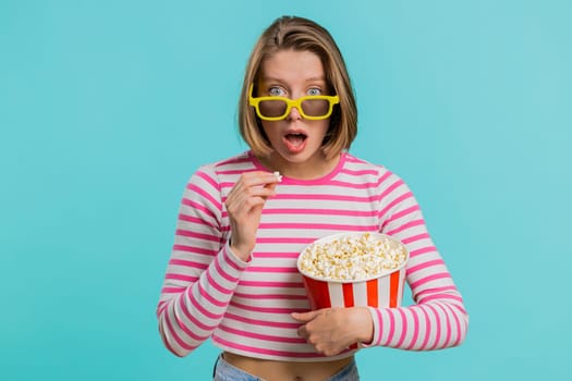 Excited young woman in 3D glasses eating popcorn and watching interesting tv serial, sport game, film, online social media movie content. Lovely girl in crop top isolated on blue studio background