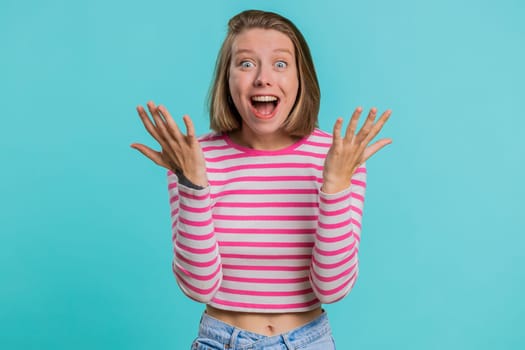 Oh my God, Wow. Excited amazed winner woman raising hands in surprise looking at camera with big eyes, shocked by sudden victory. Sincere girl in crop top isolated alone on blue studio background