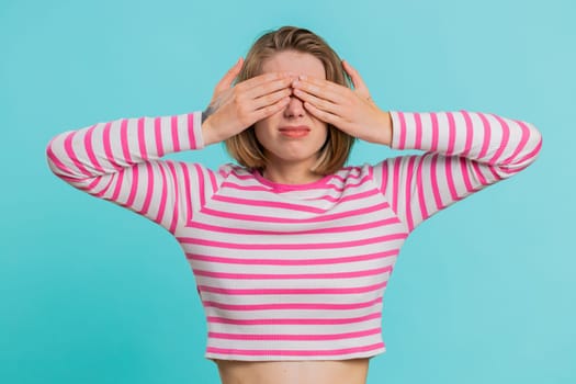 Dont want to look at this, awful. Afraid woman in crop top closing eyes with hand showing stop gesture, confused shy scared to watch. Young girl isolated on blue studio background. People lifestyles