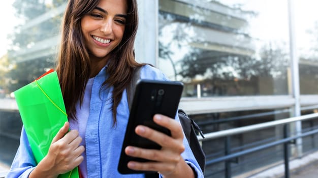 Horizontal banner image of happy young female university student looking mobile phone standing outside university building. Copy space. Education concept.