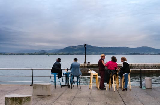 Small group of tourists sitting quietly on the terrace of a bar with the bay of Santander in the background.