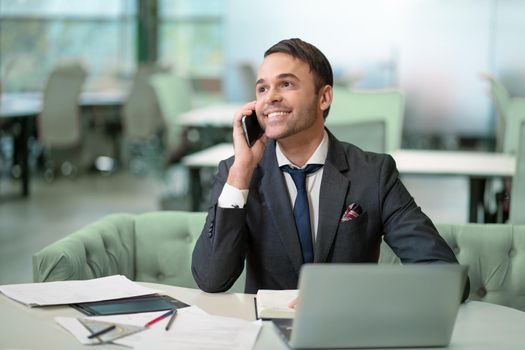 Handsome man in business suit talking on the phone working on laptop, job in progress. Cheer smiling freelancer making notes in scheduler sitting in bright coworking space.