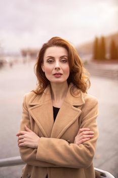 Pretty Parisian woman in an autumn beige coat beautiful hair standing outdoors posing for camera with urban city background. Vertical portrait. Retro tinted photo. 