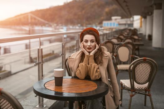 Disappointed or frustrated french young woman sitting outdoors restaurant terrace with coffee mug female fashion. Portrait of stylish young woman wearing autumn coat and red beret outdoors. 