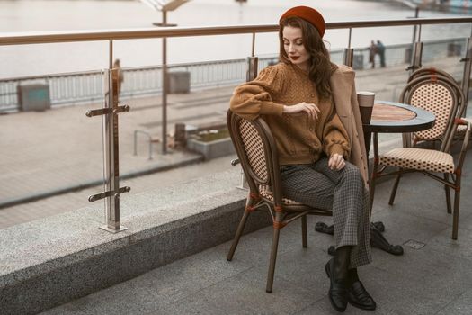 Waiting or frustrated french young woman sitting outdoors street restaurant looking frustrated down. Portrait of stylish young woman wearing autumn coat and red beret outdoors. 