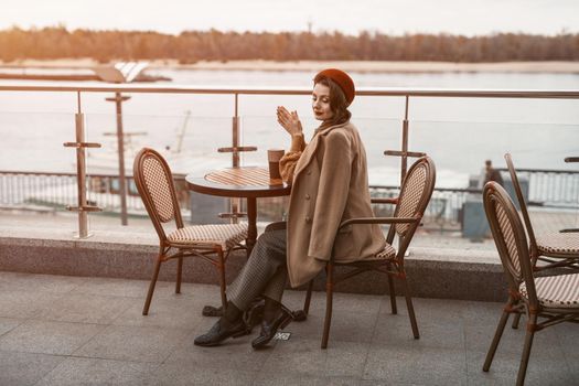 Overall plan of a thoughtful young Parisian woman with a coffee mug on the table wearing red beret sitting on terrace of restaurant or cafe. Autumn urban city on background. Tinted image. 