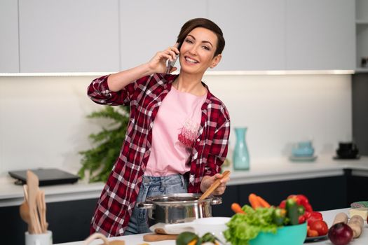 Mixing a soup or ragout while talking on the phone housewife with a short hairstyle. Cooking lunch for family in the modern kitchen. Healthy food at home. Healthy food leaving concept. 