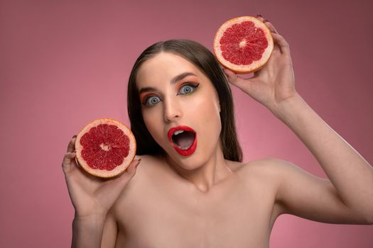 Beauty portrait of an attractive young woman holding two parts of juicy grapefruit sliced in her hands looking surprised. Charming joyful funny lady with long hair isolated on pink background.