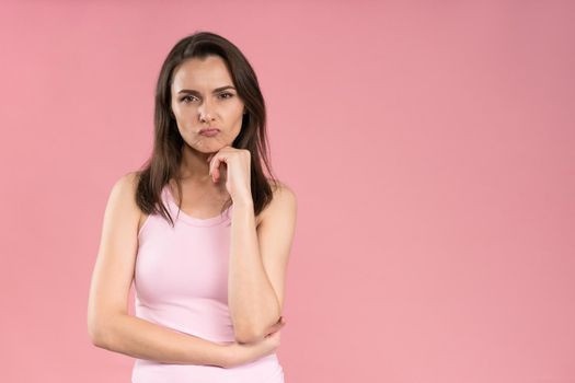 Annoyed or bothered young woman wearing pink t-shirt holding hands folded with chin leaned on one, Female half-length portrait. Human emotions, facial expression concept.