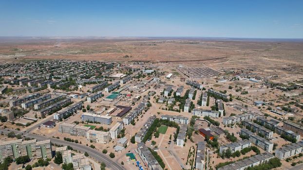 Drone view of the small town of Balkhash. The city is located on the shore of a lake. Low houses, free streets and roads. Green trees grow and there are sports grounds. There is a steppe around city