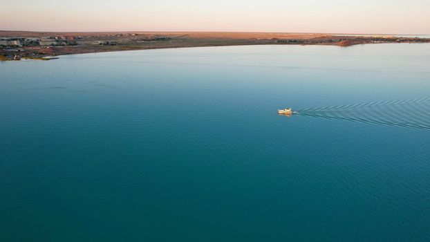 The boat sails in the bay at sunset near the city. In the distance, you can see the pipes of the plant and the orange sun. Ecology of Lake Balkhash. Catamarans float, city lights are lit. Kazakhstan