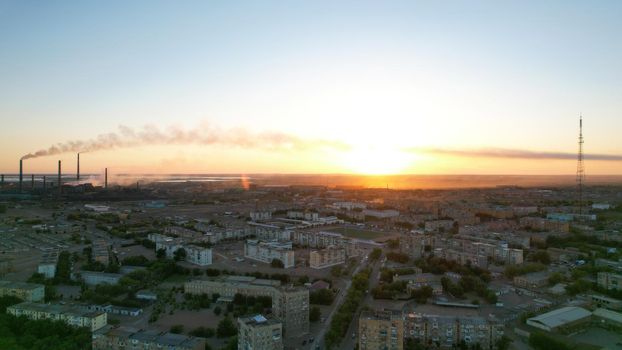 An epic sunset with a view of the smoking factory and the city. The embankment of Lake Balkhash. Low houses are standing and there are few trees. The pipes from the factory are smoking. Kazakhstan