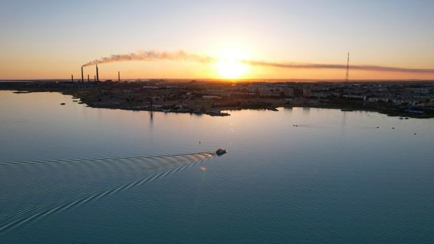 The boat sails in the bay at sunset near the city. In the distance, you can see the pipes of the plant and the orange sun. Ecology of Lake Balkhash. Catamarans float, city lights are lit. Kazakhstan