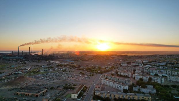 An epic sunset with a view of the smoking factory and the city. The embankment of Lake Balkhash. Low houses are standing and there are few trees. The pipes from the factory are smoking. Kazakhstan