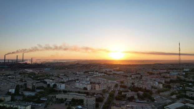 An epic sunset with a view of the smoking factory and the city. The embankment of Lake Balkhash. Low houses are standing and there are few trees. The pipes from the factory are smoking. Kazakhstan