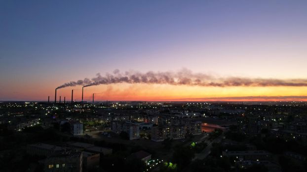 An epic sunset with a view of the smoking factory and the city. The embankment of Lake Balkhash. Low houses are standing and there are few trees. The pipes from the factory are smoking. Kazakhstan